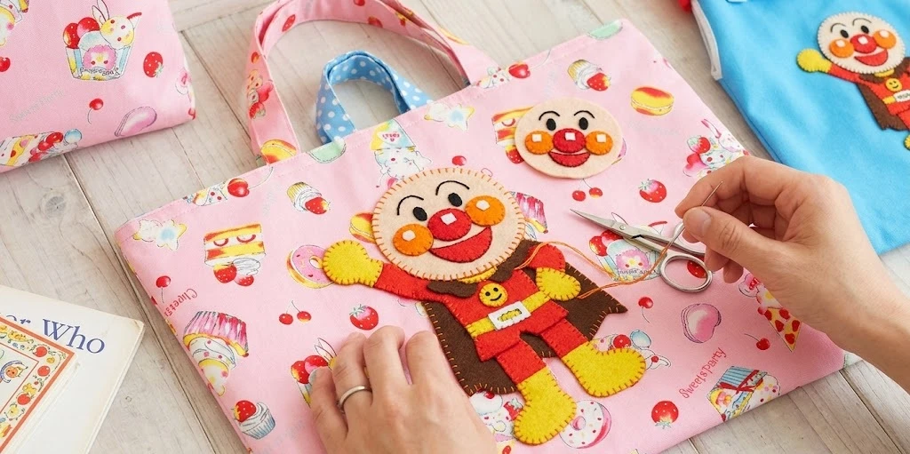 お菓子柄の手作りレッスンバッグに、フェルトで作ったアンパンマンのマスコットワッペンを縫い付けている手元の様子。